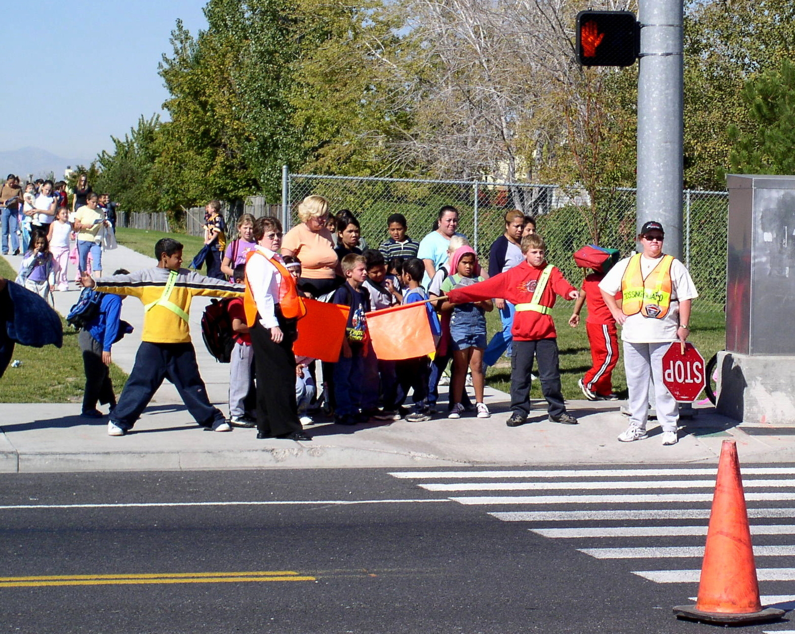 kids waiting to cross the street beind directed by a crossing guard.